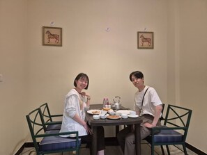 Dos personas sentadas a una mesa con platos, tazas y una tetera, en una habitación con sillas e imágenes enmarcadas en la pared.