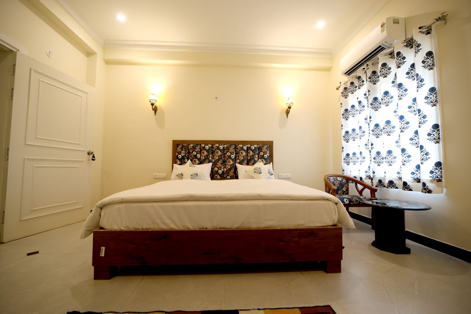 Un dormitorio con cama, lámparas de noche, una ventana con cortinas estampadas y una silla.