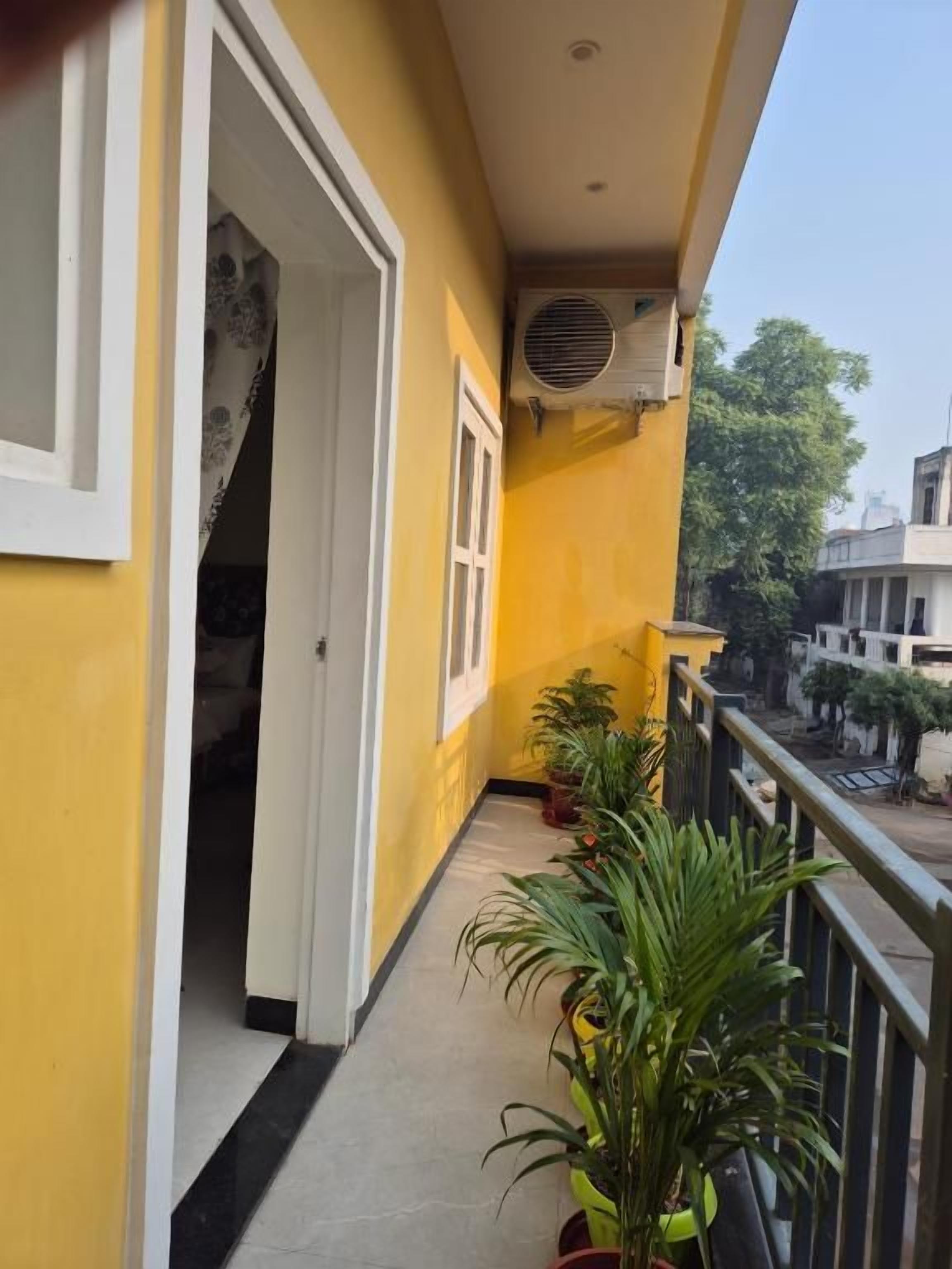 A narrow balcony with a yellow wall, a door leading inside, and potted plants.