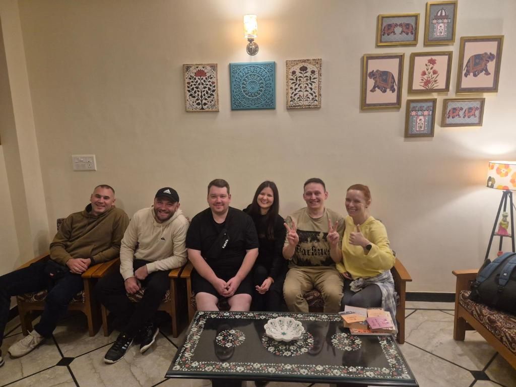 A group of people seated in a room with a table in the center, a sofa, and wall decorations including framed pictures and a wall sconce.