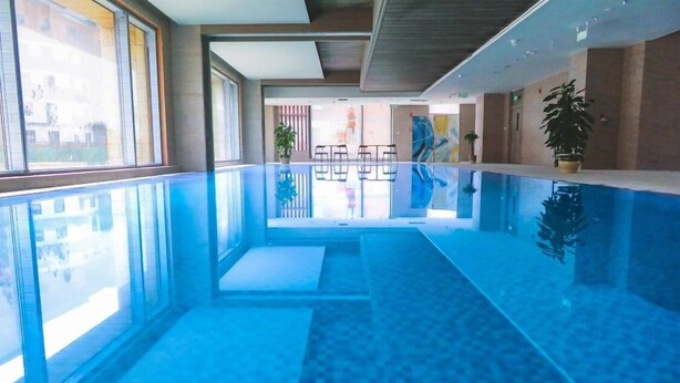 A modern indoor swimming pool with clear glass walls, a tiled floor, and potted plants. - Courtyard By Marriott Chongli (Zhangjiakou)