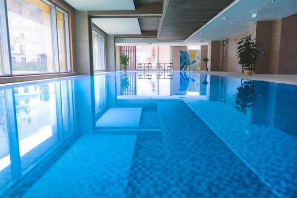 Une piscine intérieure moderne avec des parois en verre transparent, un sol carrelé et des plantes en pot.