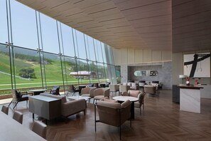 Un hall d’hôtel moderne doté d’une grande paroi vitrée donnant sur la campagne. On y trouve plusieurs espaces de détente avec des tables et des chaises.