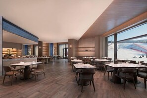 Un vaste espace dédié à la salle à manger, avec de nombreuses tables et chaises, de grandes fenêtres offrant une vue sur un paysage enneigé et un aménagement intérieur moderne.