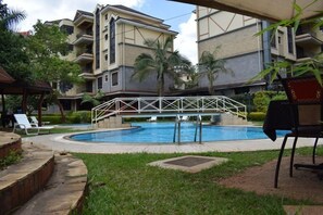 A swimming pool surrounded by a white fence, with a covered seating area and a view of multi-story buildings in the background. - Kengen RBS Gardens Apartments (Nairobi)