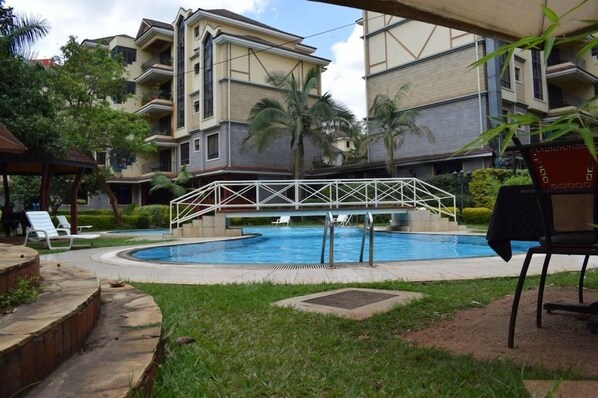 A swimming pool surrounded by a white fence, with a covered seating area and a view of multi-story buildings in the background. - Kengen RBS Gardens Apartments (Nairobi)