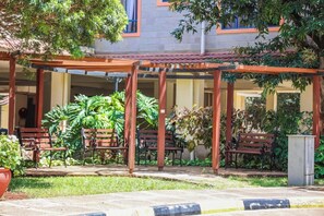 An outdoor seating area with wooden benches and a pergola structure, surrounded by greenery and a building with a tiled roof. - Kengen RBS Gardens Apartments (Nairobi)