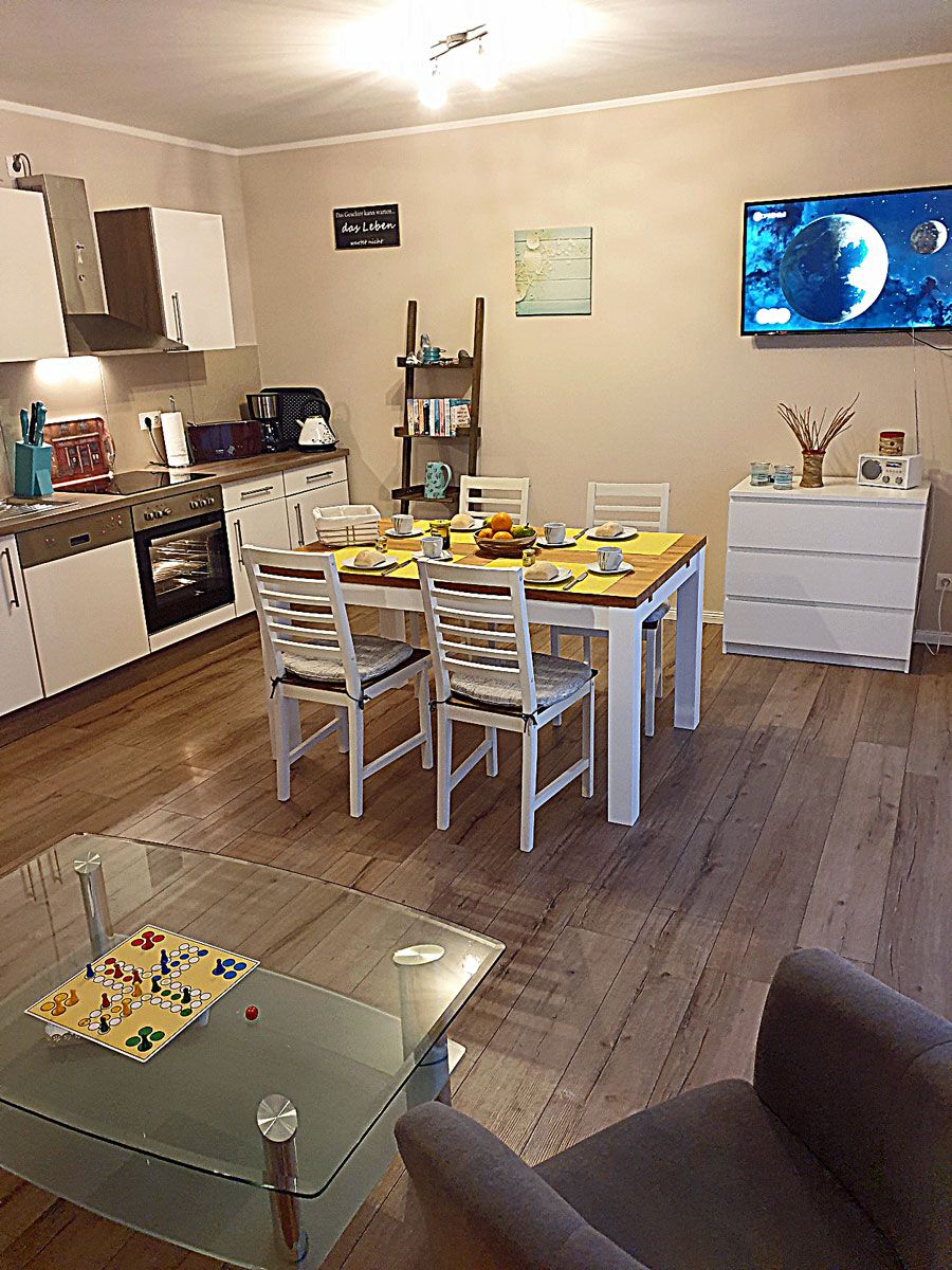 Una cocina moderna con armarios blancos, una mesa de comedor con sillas, un televisor en la pared y una mesa de centro de vidrio con un juego de mesa.