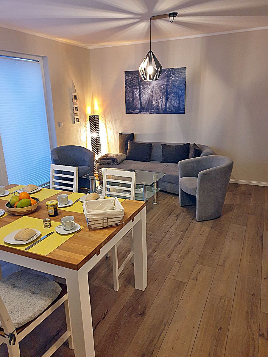 Ein Wohnzimmer mit Esstisch für zwei Personen, einem grauen Sofa und einer modernen Pendelleuchte.