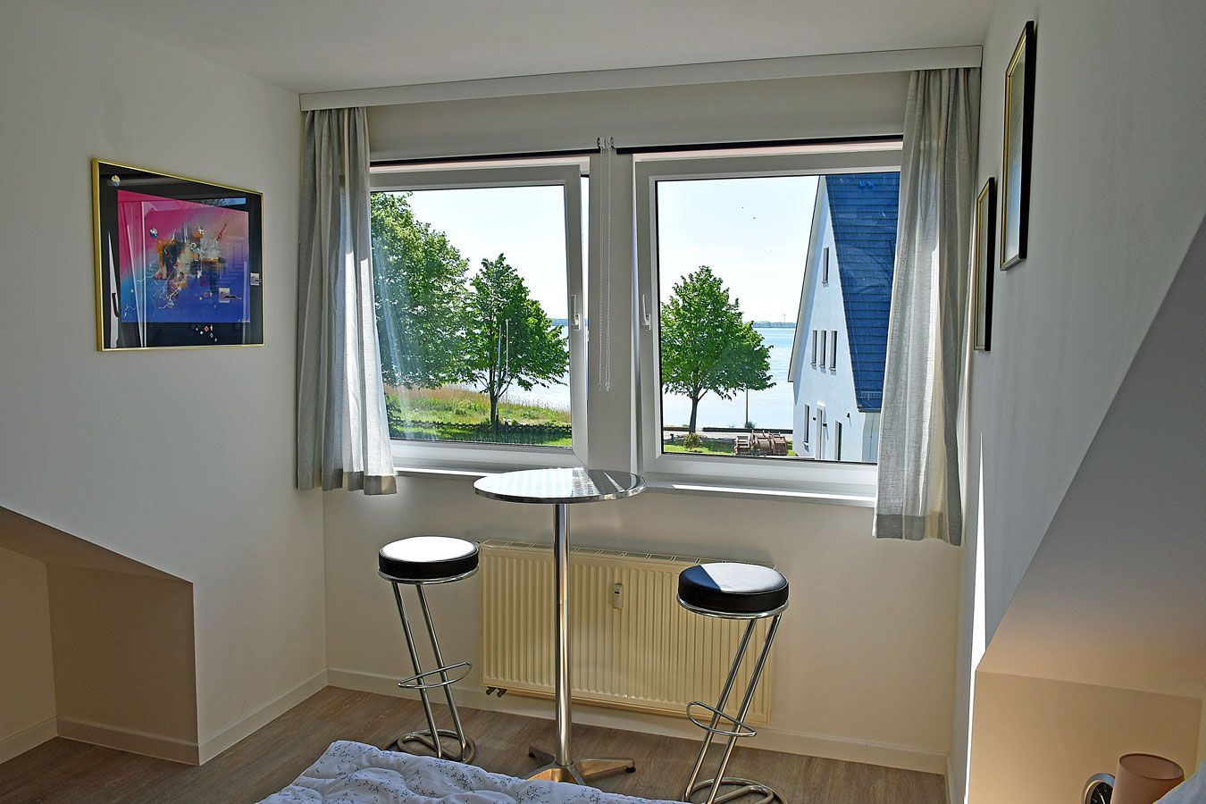 Een kamer met een groot raam, een radiator, twee barkrukken en een ingelijste foto aan de muur.