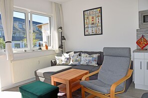 A cozy living room with a sofa, a wooden table, a green ottoman, a radiator, a window with curtains, a wall-mounted lamp, and a framed picture of lighthouses. - Modern vacation rental with ocean views, just 60 meters from the water, 2 bedrooms, Wi-Fi, pets allowed (Dranske)