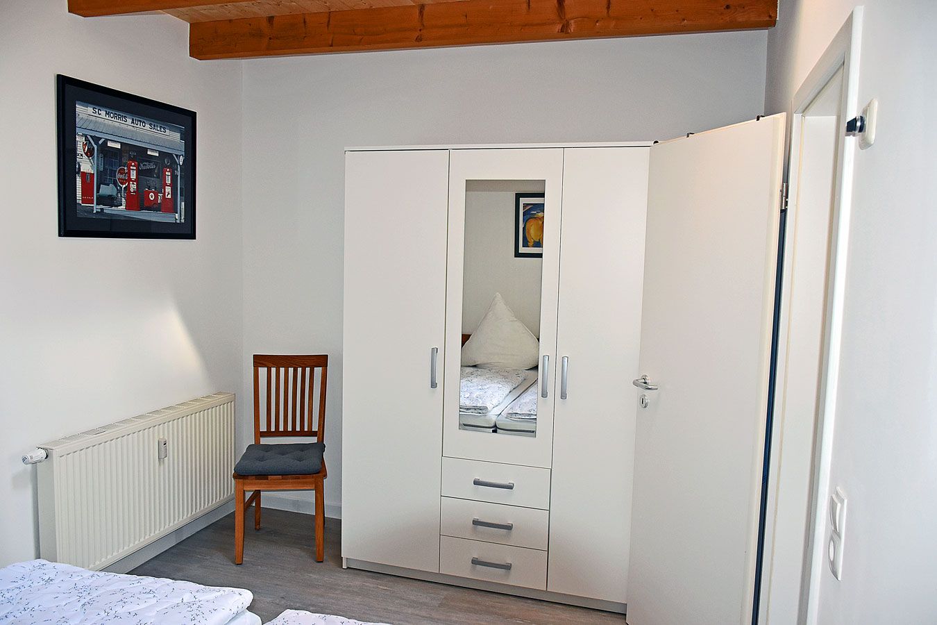 Een slaapkamer met een witte garderobe, een houten stoel, een radiator en een ingelijste foto aan de muur.