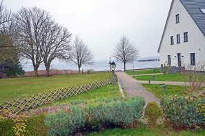 A pathway leading to a building with a white facade and multiple windows, surrounded by a grassy area with trees and a body of water in the background. - Vacation rental with a view of the water, 2 bedrooms, just 60 meters from the water, Wi-Fi (Dranske)