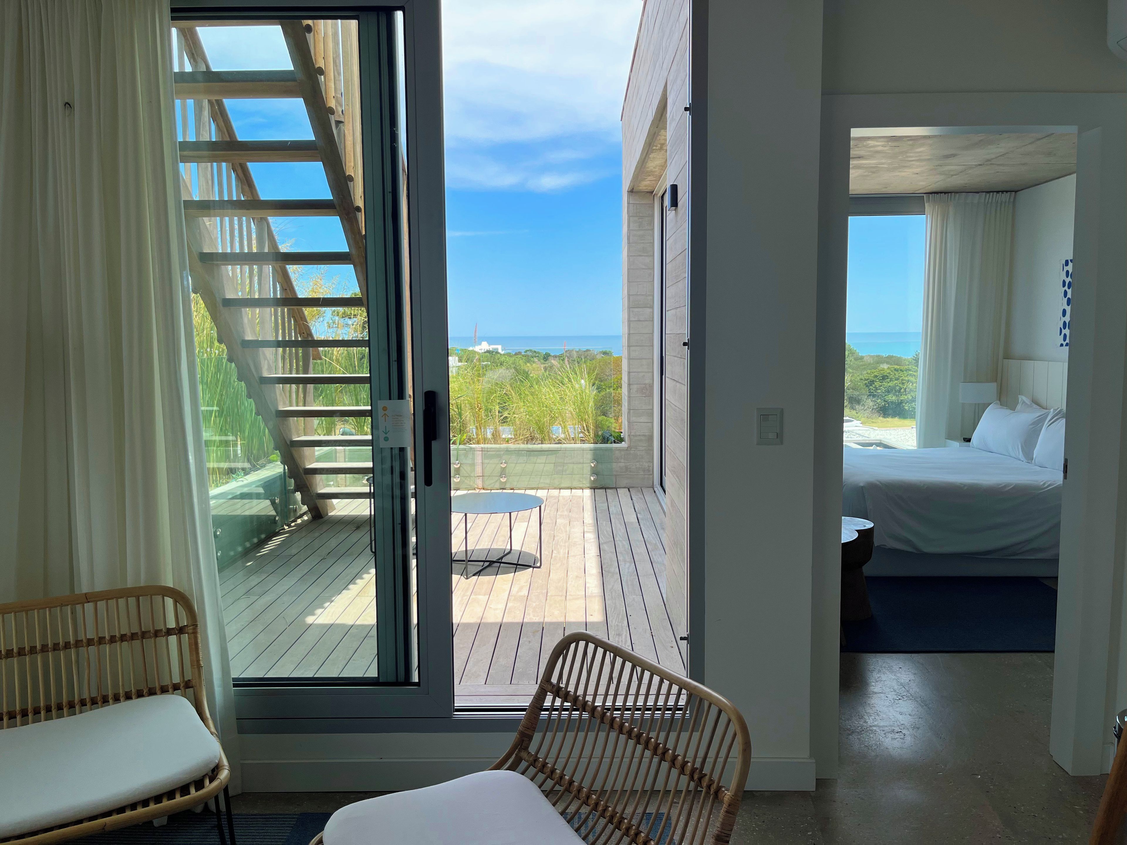 A room with a bed, a wicker chair, and a view of the ocean through a sliding door.