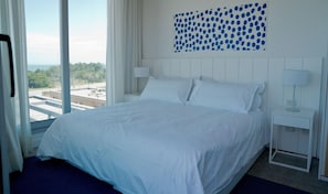 A hotel room with a large bed, a nightstand, a lamp, and a view of the cityscape through the window. - Casa Grande Hotel & Beach Club (José Ignacio)