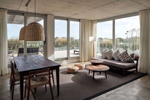 Ruime kamer met een eettafel, stoelen, een bank en poefs. Grote ramen bieden uitzicht op het omliggende landschap.