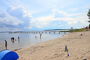 Ein Strand mit einem Steg, der sich ins Wasser erstreckt, Menschen, die am Strand entlanggehen und sitzen, und einem aufgebauten Zelt.