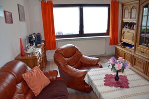 Olohuoneessa on nahkasohva, nahkainen nojatuoli, puinen kaappi ja ikkuna, jossa on oranssit verhot.