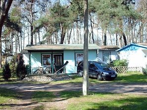 A two-story building with a porch, a car parked in front, and a forested background. - Bungalow right on the sandy beach of the Baltic Sea, sleeps up to 2 people, with 1 bedroom (Juliusruh)