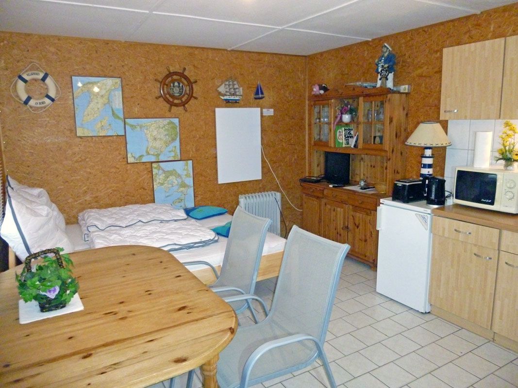 Et lille værelse med seng, et spisebord med stole, et tekøkken med mikroovn og et træskab.