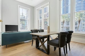 A modern living room with a dining table, chairs, a TV, and large windows.