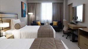 Quarto de hotel com duas camas, uma escrivaninha, uma cadeira, um abajur e uma janela grande com cortinas.