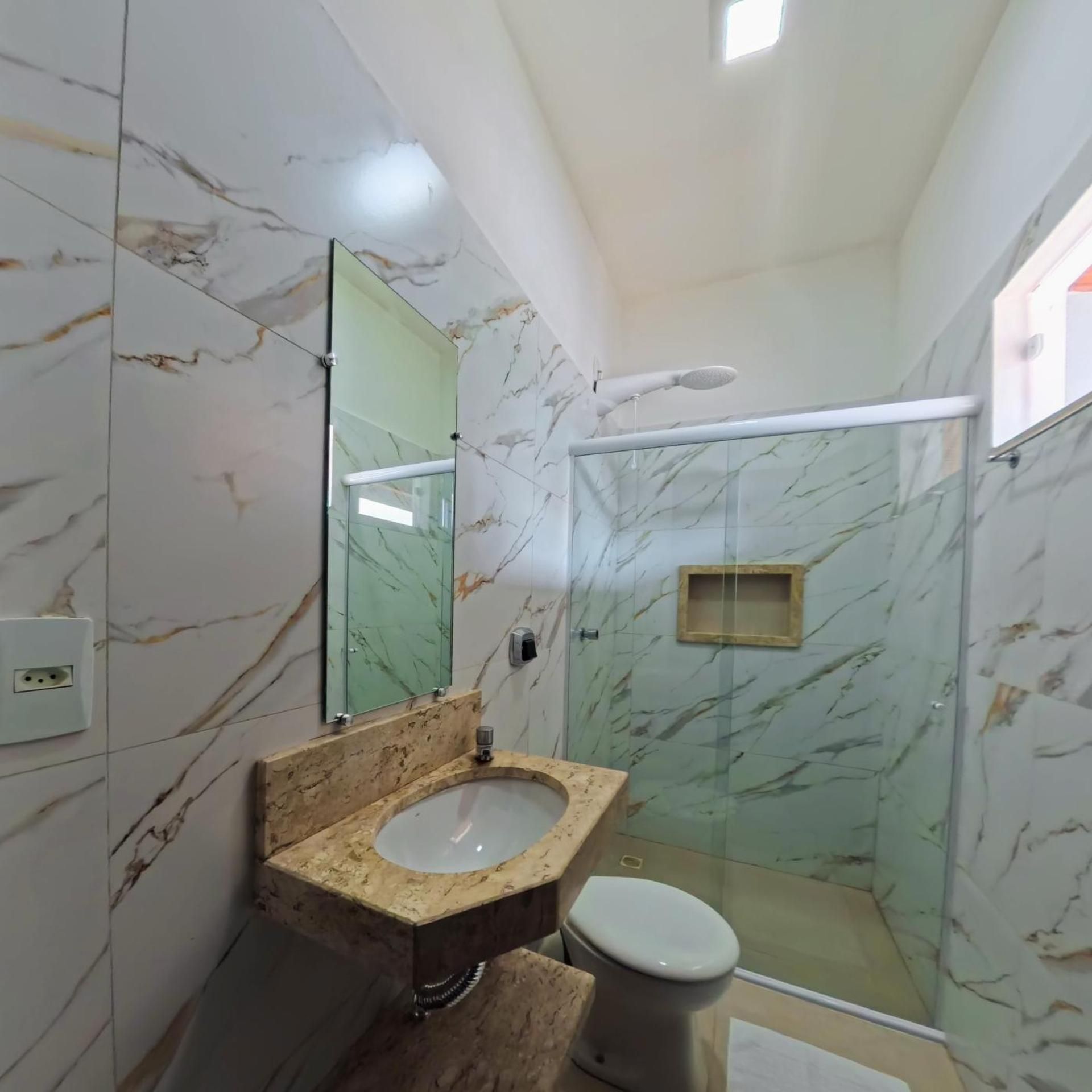 Um banheiro com parede de mármore, box de vidro, pia com bancada de mármore e vaso sanitário.