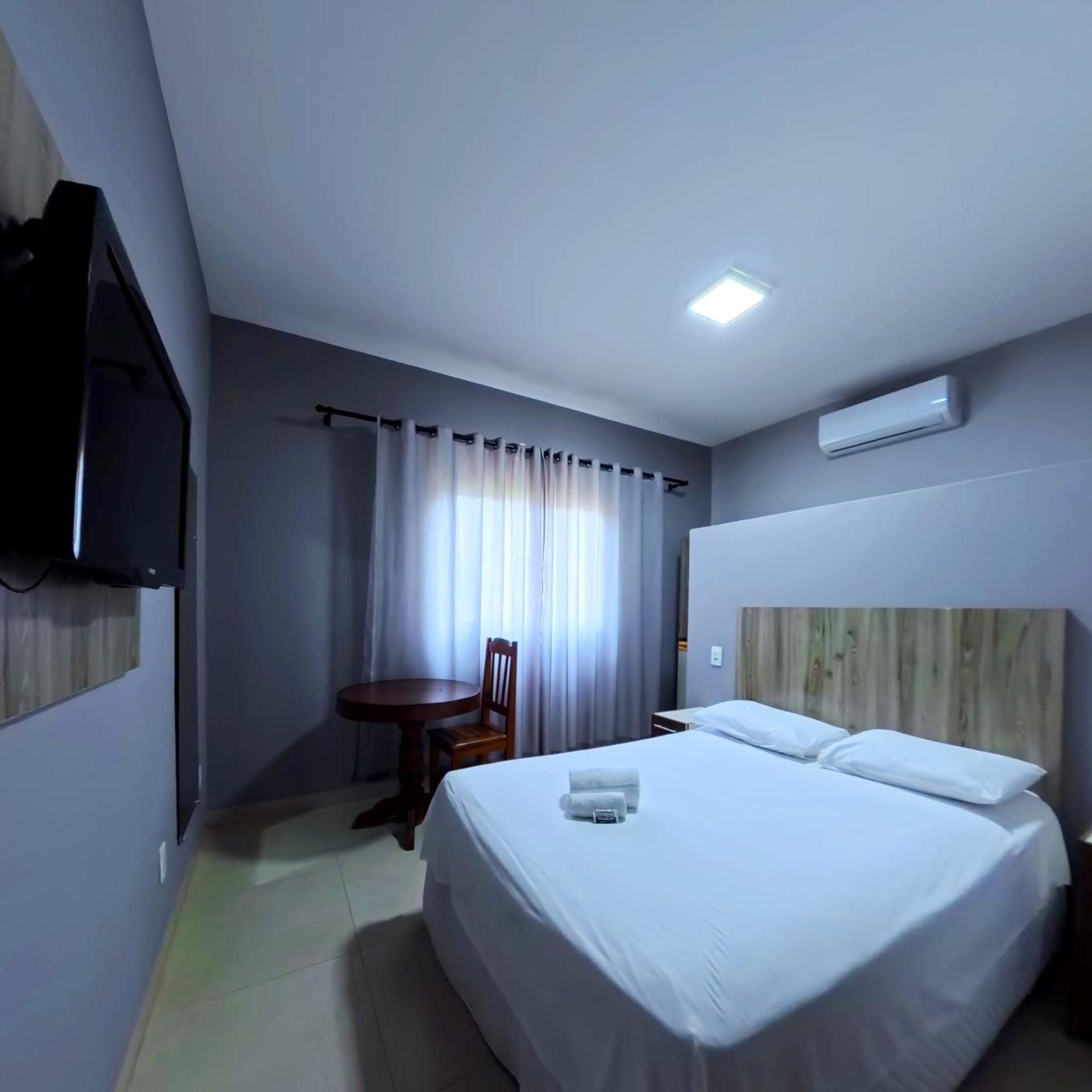 Quarto de hotel com cama, uma cadeira, uma mesinha, TV fixada na parede e ar condicionado.