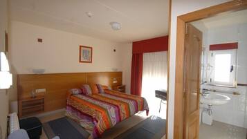 Habitación de hotel con cama, mesitas de noche, un escritorio, una silla, baño con lavamanos y espejo, y una puerta que da a otra habitación.