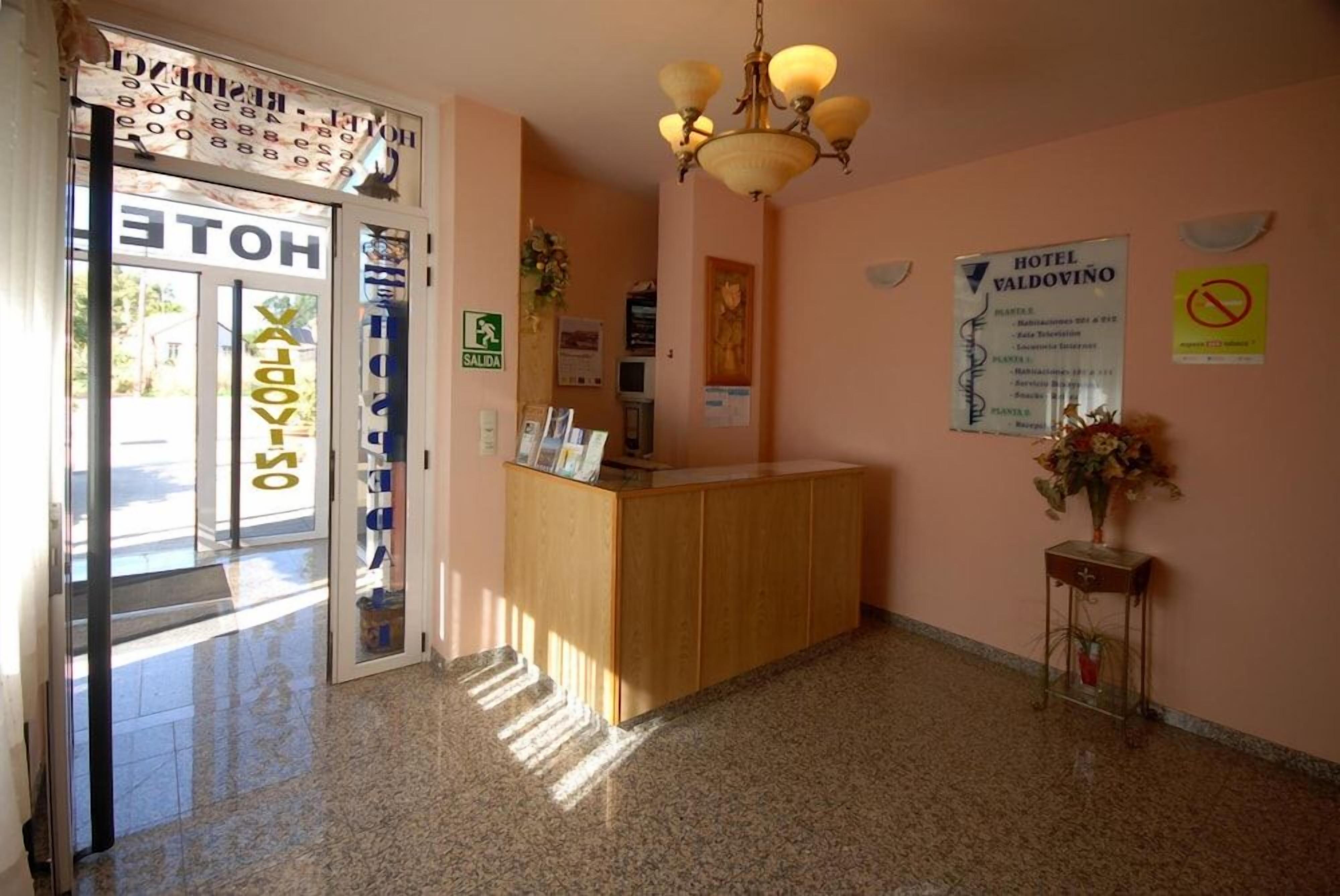 Zona de recepción de un hotel con un mostrador de madera, una araña y varios carteles informativos en las paredes.