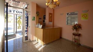 Zona de recepción de un hotel con un mostrador de madera, una araña y varios carteles informativos en las paredes.