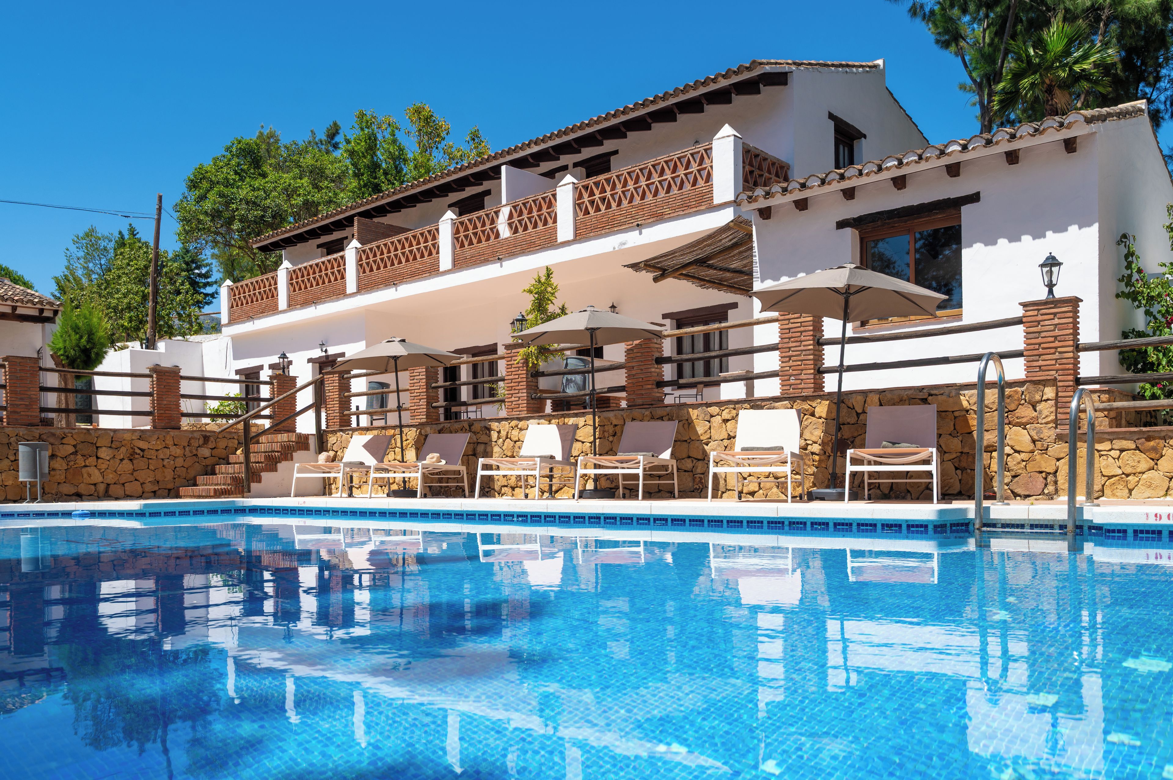 A poolside area with lounge chairs and umbrellas, a stone wall, and a building with a tiled roof and balconies.