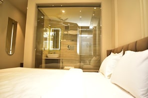 A hotel room with a bed, a bathroom visible through a glass door, and a ceiling fan. - Hotel Cullinan By Qotel (New Delhi)