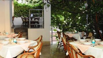 Ein Essbereich im Freien mit Tischen und Stühlen, der von einer mit Weinreben bewachsenen Pergola überdacht wird.