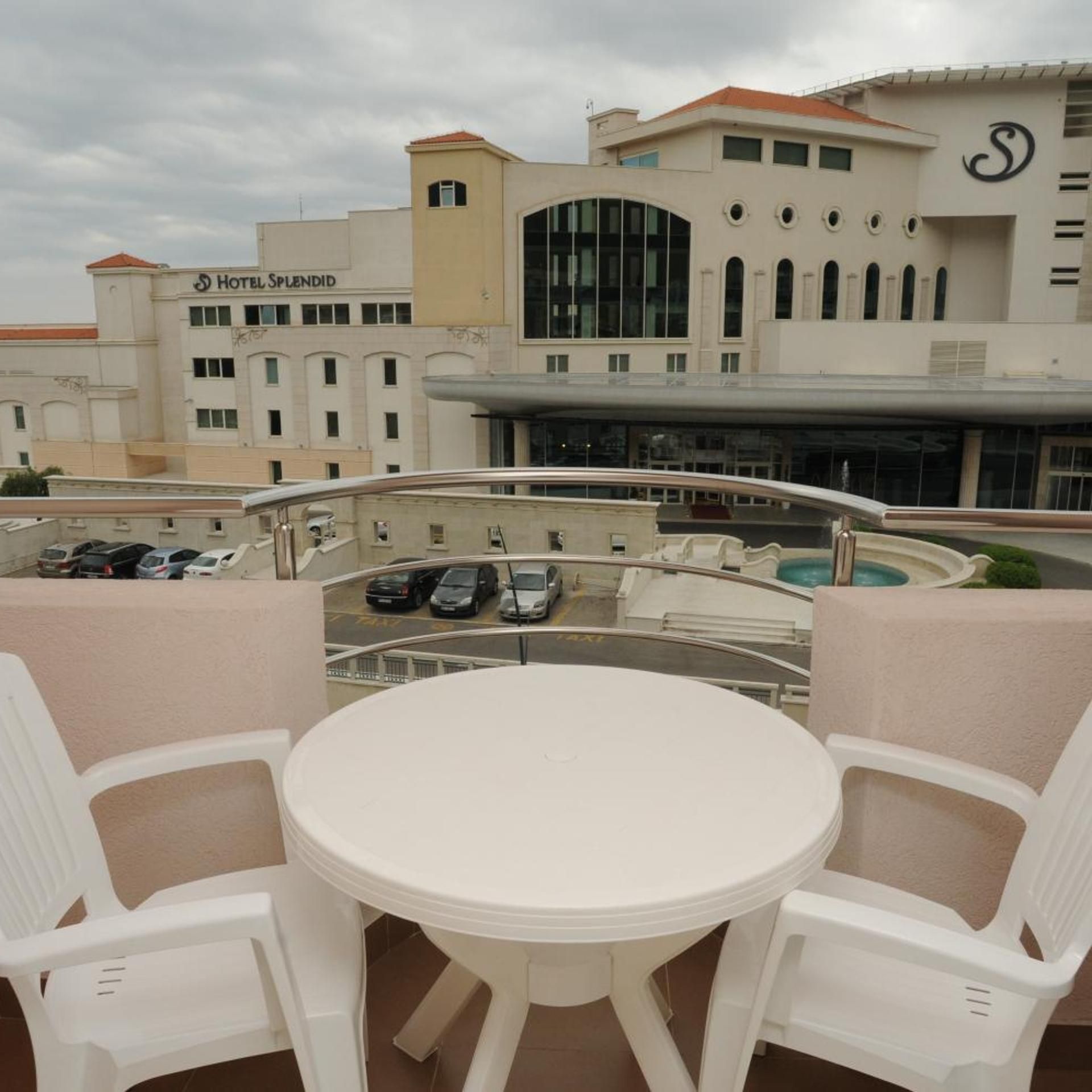 Eine Dachterrasse mit rundem Tisch und Stühlen, mit Blick auf einen Parkplatz und ein Hotelgebäude mit der Aufschrift 'HOTEL SPLENDID'.