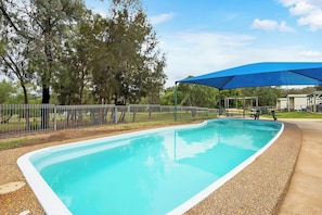 Una piscina rectangular con una lona azul por encima, rodeada por una valla, árboles y un edificio al fondo.