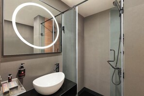 Un baño moderno con un lavamanos blanco, un cabezal de ducha de mano y un espejo grande con luz circular.