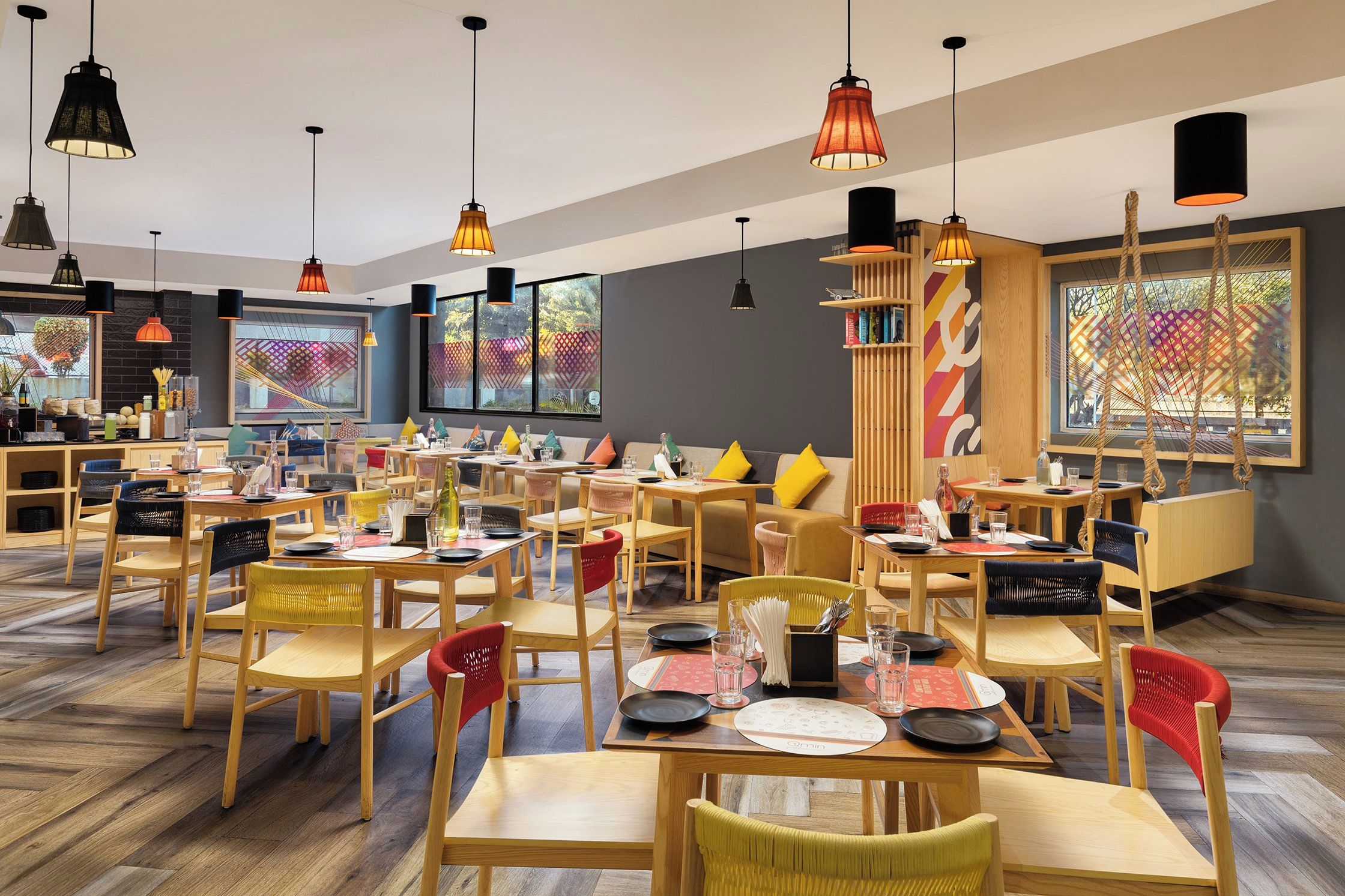 A restaurant with multiple dining tables and chairs, overhead pendant lighting, and large windows with artwork.