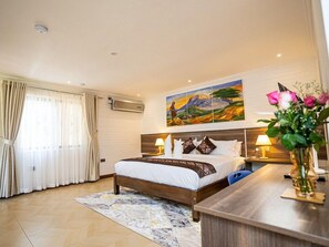 A hotel room with a bed, bedside lamps, a painting above the bed, a window with curtains, and a vase with flowers on a table. - Light Villas (Nairobi)