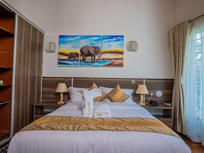 A hotel room with a large bed, two bedside tables, a painting of elephants, and a window with curtains. - Light Villas (Nairobi)