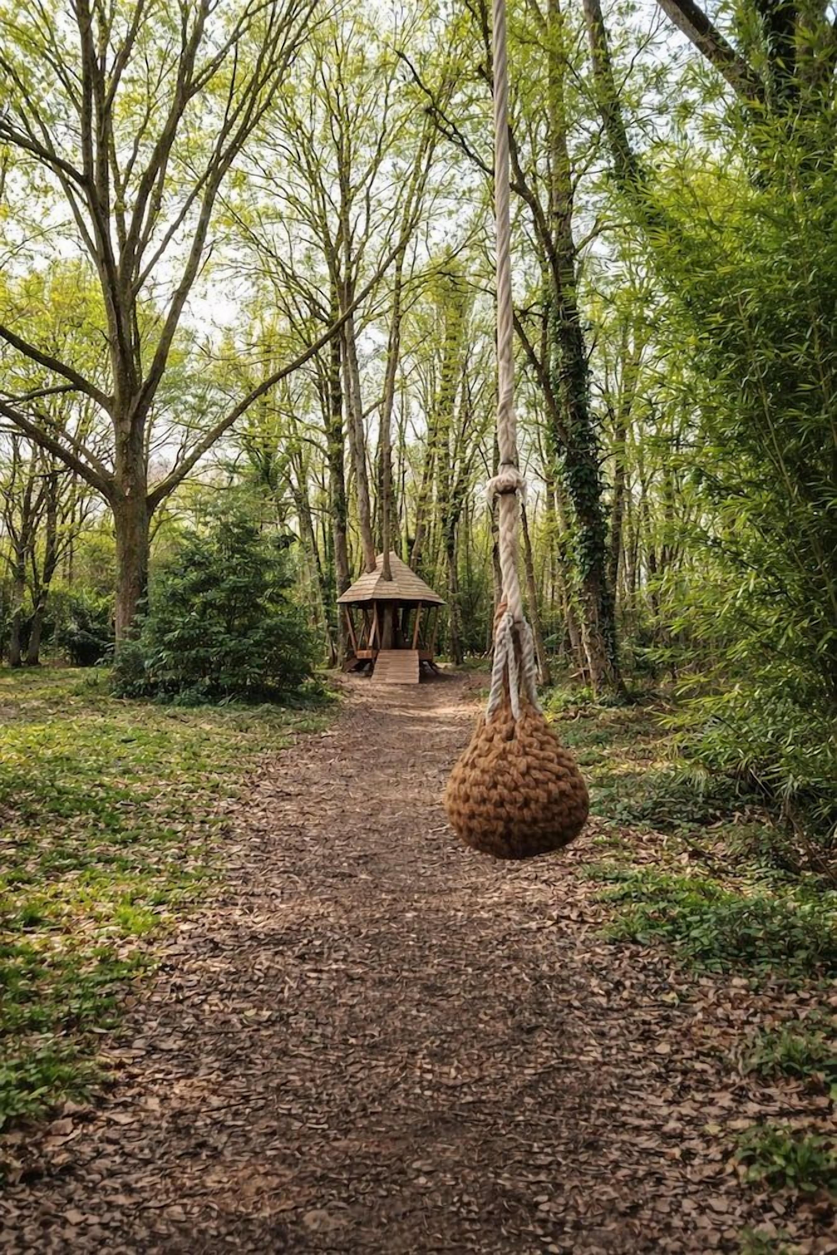 Un sentier forestier mène à une construction rustique en bois, à laquelle est suspendue une balançoire faite de corde, accrochée à un arbre.