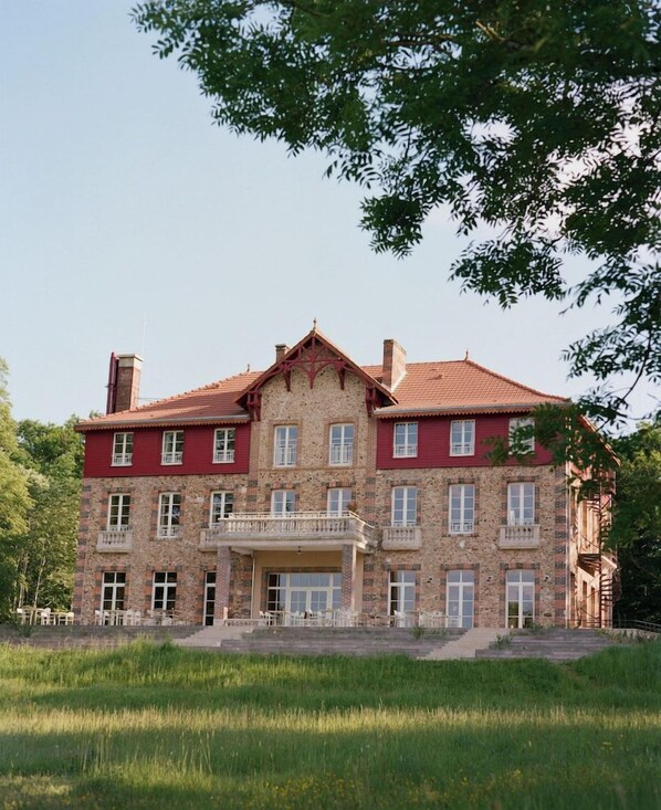Un grand bâtiment en pierre, doté de nombreuses fenêtres et de balcons, entouré d’une zone verdoyante.