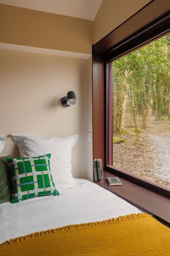 Un dormitorio con un ventanal grande, una cama con ropa de cama blanca, un almohada verde y blanca, un plaid amarillo y una lámpara de lectura fijada a la pared.