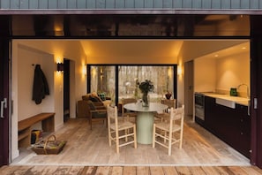An open-plan living area with a dining table, chairs, a sofa, and a kitchen area visible through the glass doors.