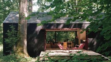 Una cabaña rústica con fachada de madera, un porche cubierto con sillas y vistas al bosque circundante.
