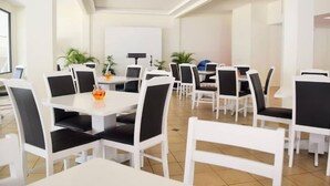 Uma sala de jantar espaçosa com mesas e cadeiras brancas, algumas com almofadas pretas.