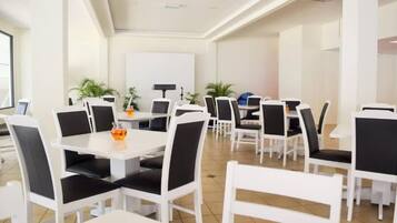 A modern dining area with white tables and chairs, potted plants, and a whiteboard on the wall.