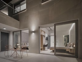 Un interior moderno con un vidrio que da a un dormitorio, una zona de estar con sillas y una escalera.