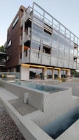 Un edificio moderno de varios pisos con fachada de vidrio, piscina en la azotea y un área ajardinada con fuente de agua.
