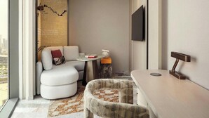 Quarto de hotel moderno com sofá, mesa redonda pequena, cadeira, TV fixada na parede e vista para a cidade.